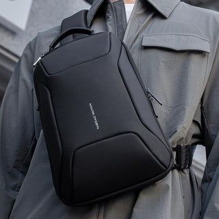 A black crossbody backpack with a top handle and front zip pocket, worn across the chest of a person in a gray jacket.