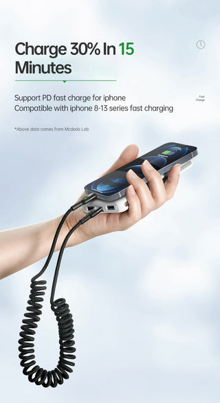 Hand holding a phone with a coiled charging cable, on a light gray background.